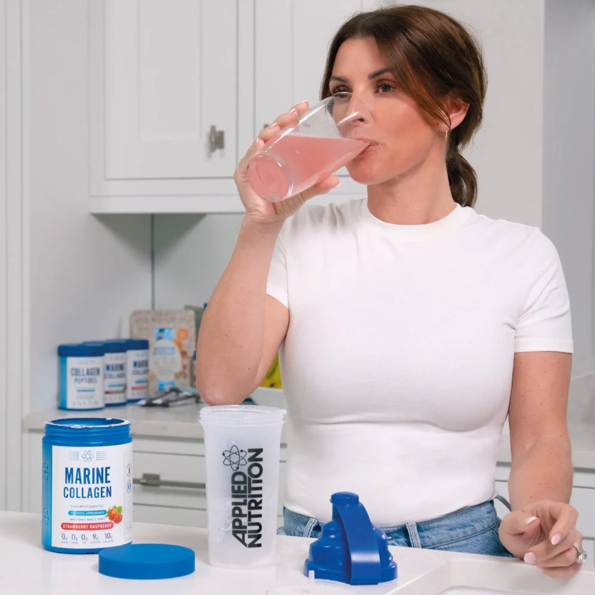 Woman drinking a pink supplement shake with Applied Nutrition products on a kitchen counter.